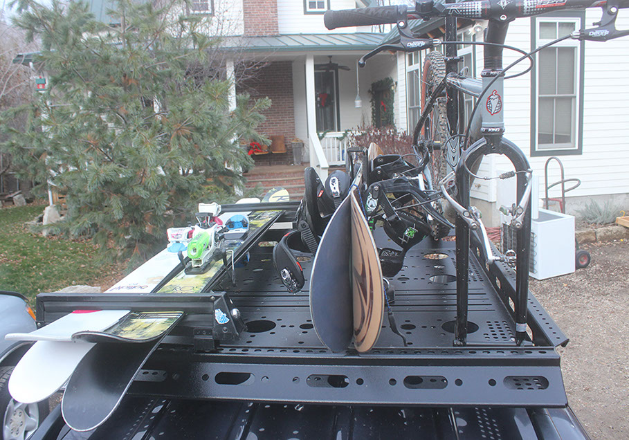 two pair of skis, two snowboards and a mountain bike attached to a small black Revrack tray atop a vehicle in front of a white house