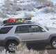 profile of a passing gold nissan pathfinder with a small revrack tray on top and winter gear including snowboards with a snowy background
