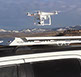 profile view of a white drone hovering above a small white revrack tray atop a white honda c r v with a foothill background